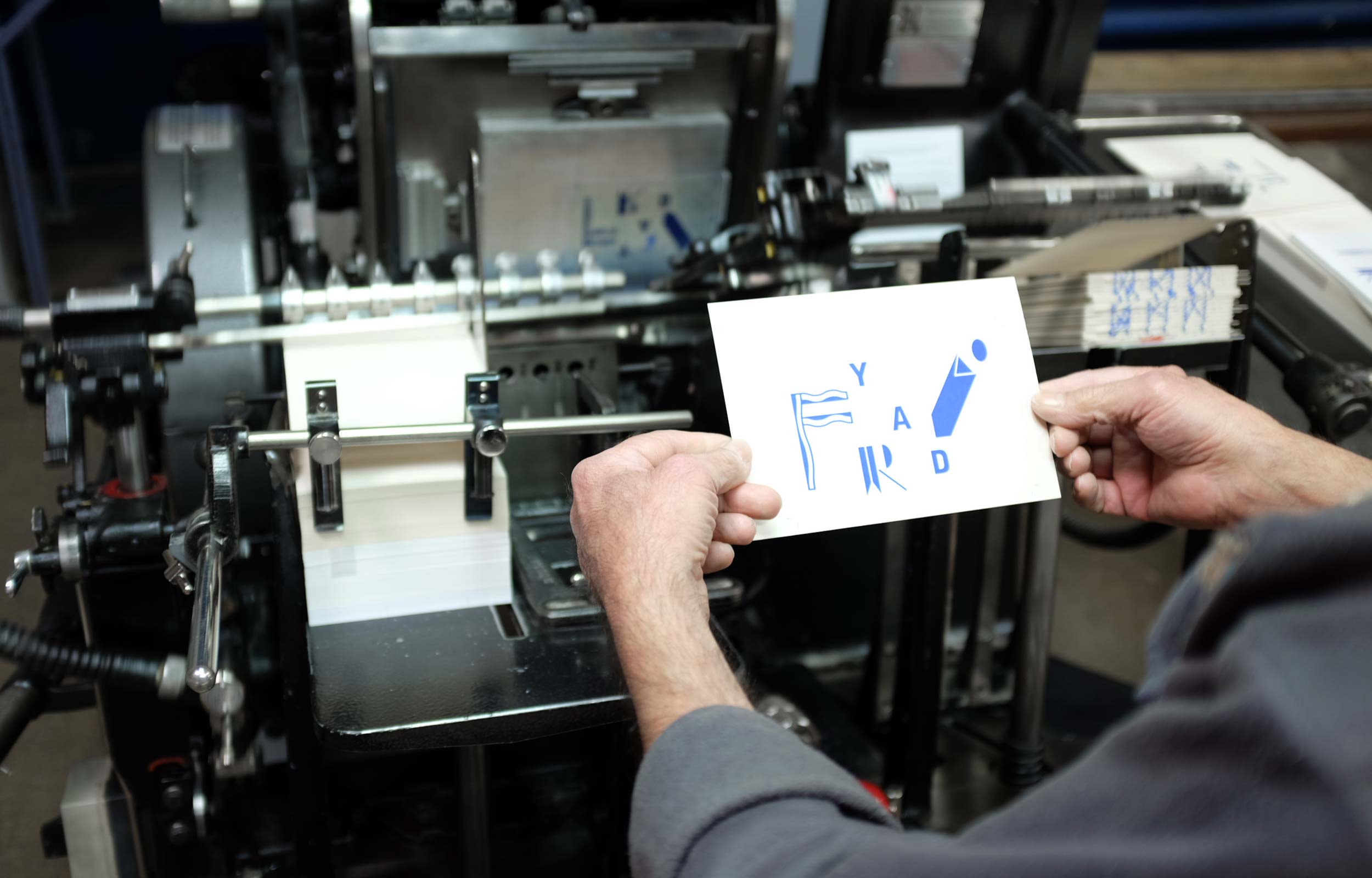 Person holding a freshly printed blue graphic card beside a printing press