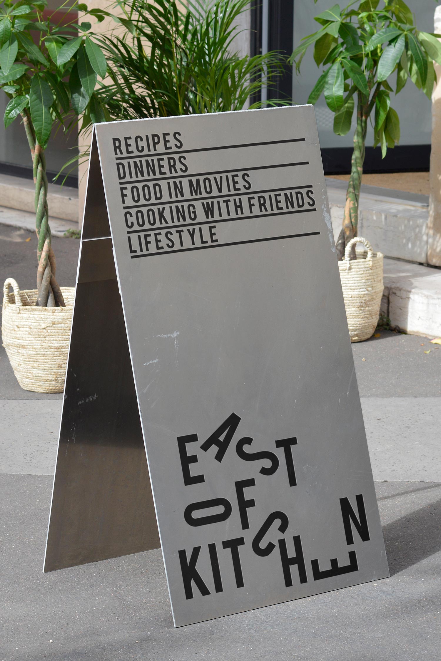 Freestanding East of Kitchen recipe sign beside potted plants