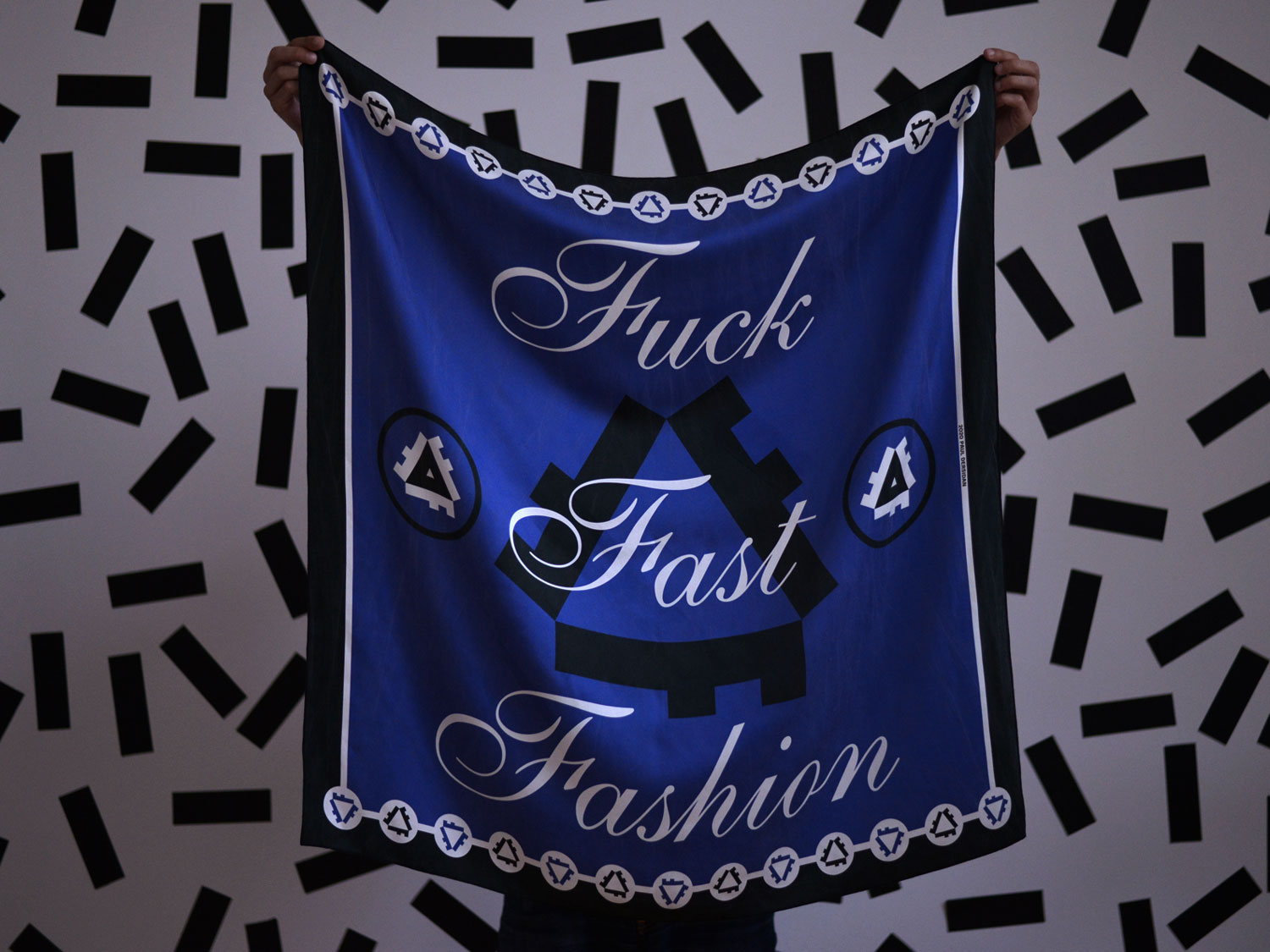 Person holding up a blue Fuck Fast Fashion scarf against a black patterned wall