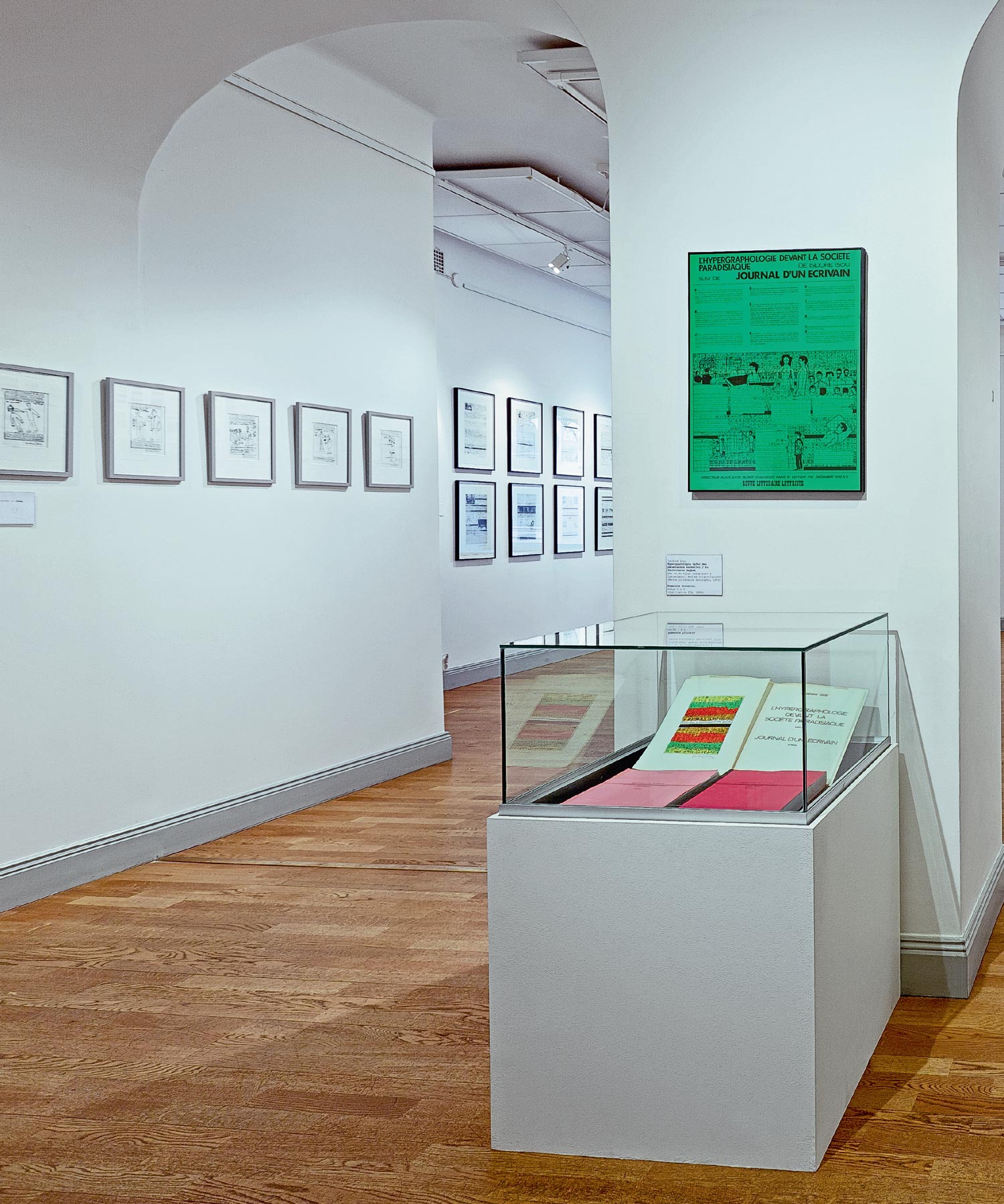 Gallery room with framed works, a green wall poster and glass display plinth