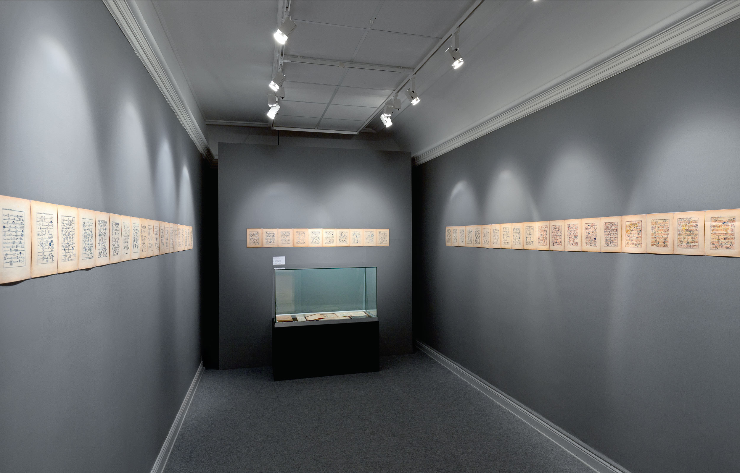 Dark-walled gallery room with framed works displayed in a long horizontal row