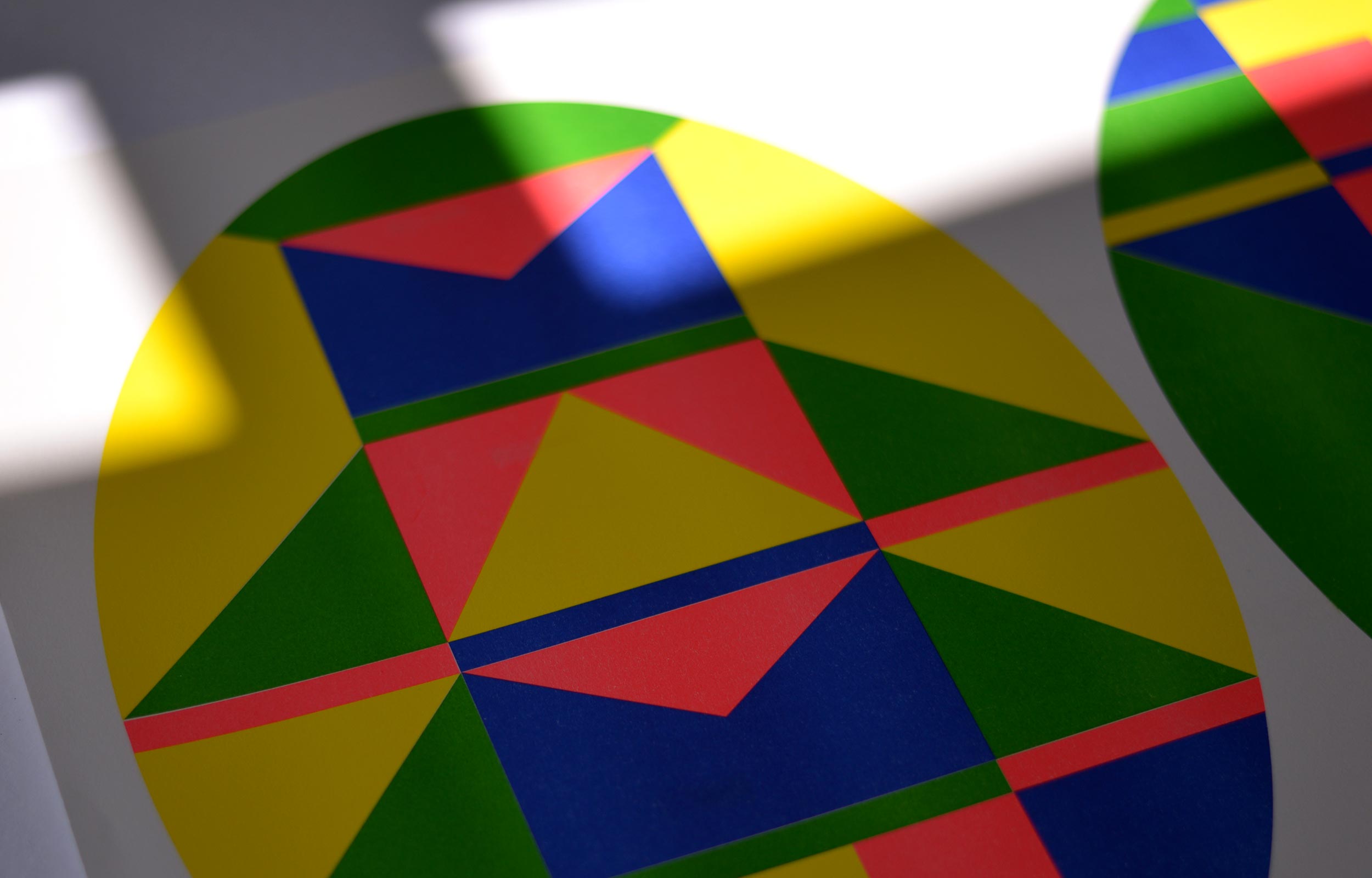 Close-up of a colourful oval geometric print on white paper, illuminated by a broad band of sunlight