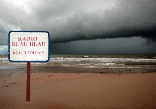 Radio Blau Blau Beach Edition sign on a post beside a cloudy shoreline